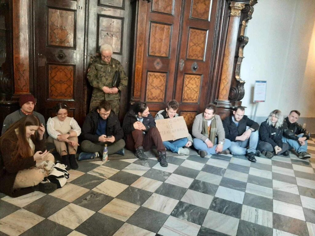 Grupa studentów siedzi na podłodze pod drzwiami, nad nimi stoi umundurowany mężczyzna